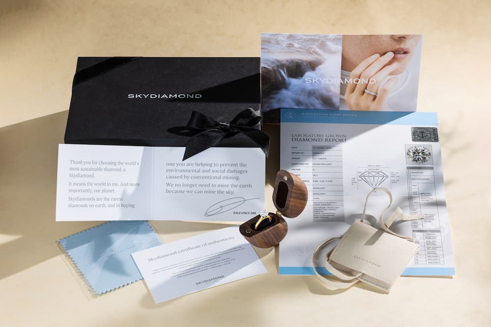A styled flat-lay of a diamond engagement set presentation including a black branded box with a black ribbon, printed cards, a diamond report, a ring box, a ring, and a small wooden ring stand on a warm beige background.